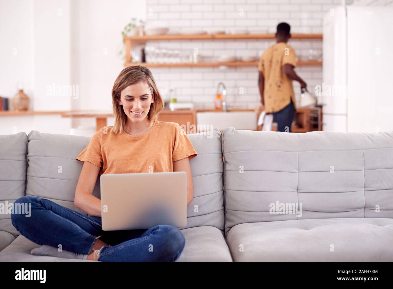 Donna seduta rilassante sul divano di casa utilizzando un computer portatile con uomo in cucina dietro Foto Stock