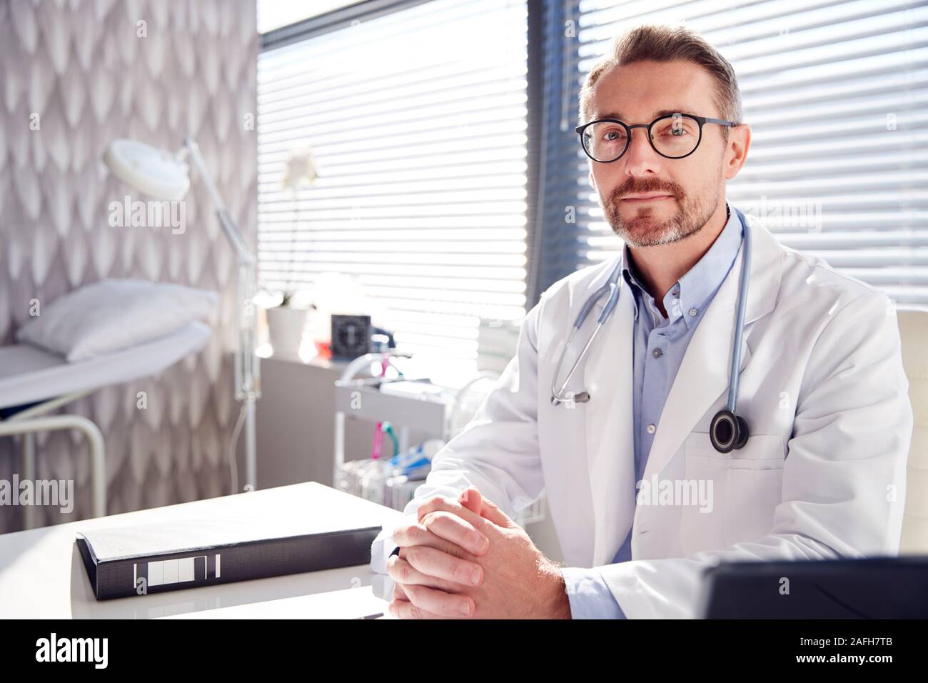 Ritratto di sorridente medico maschio indossa mantello bianco con uno stetoscopio seduto alla scrivania in ufficio Foto Stock
