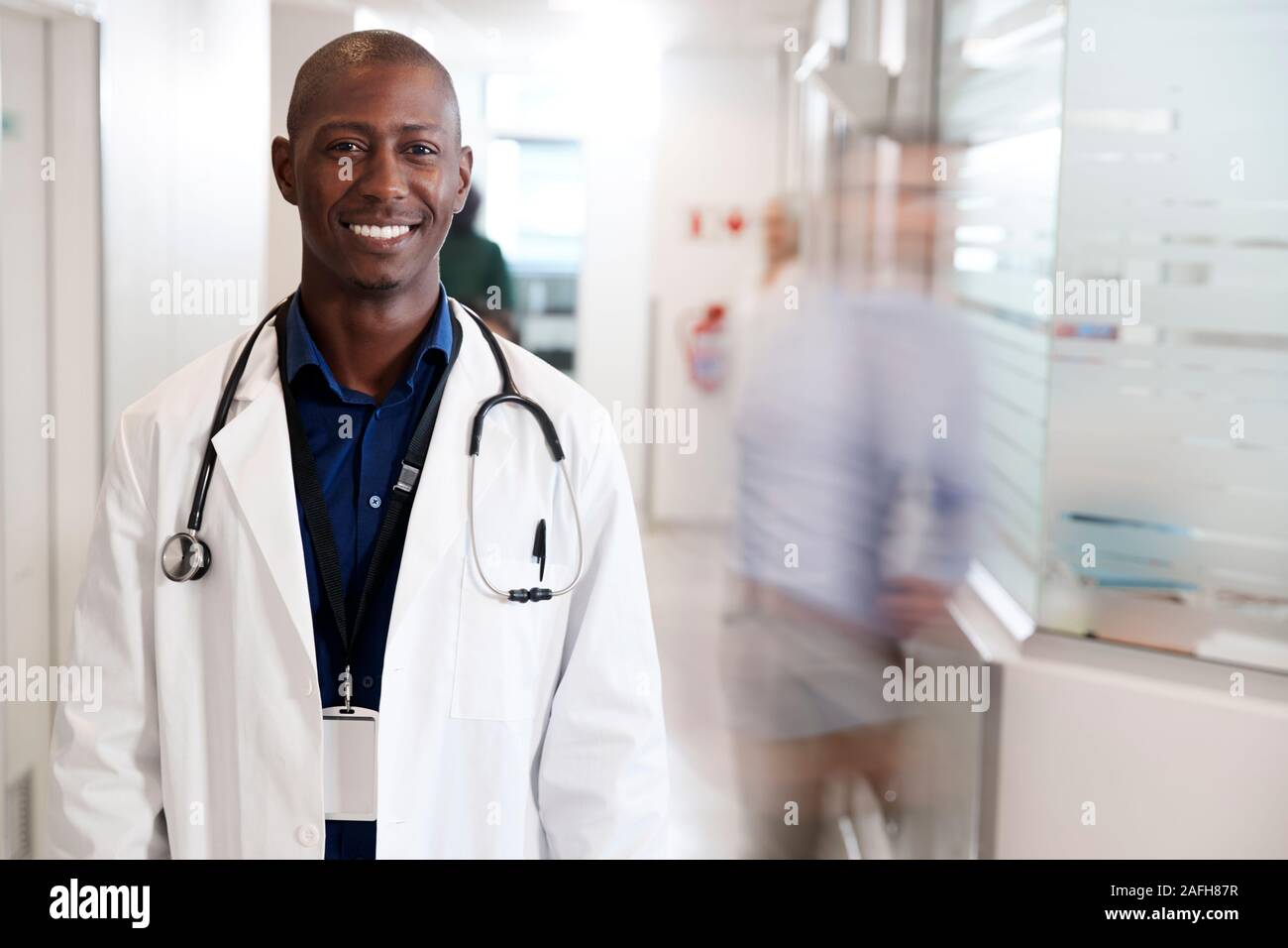 Ritratto di sorridente medico maschio indossa mantello bianco con uno stetoscopio in ospedale occupato corridoio Foto Stock