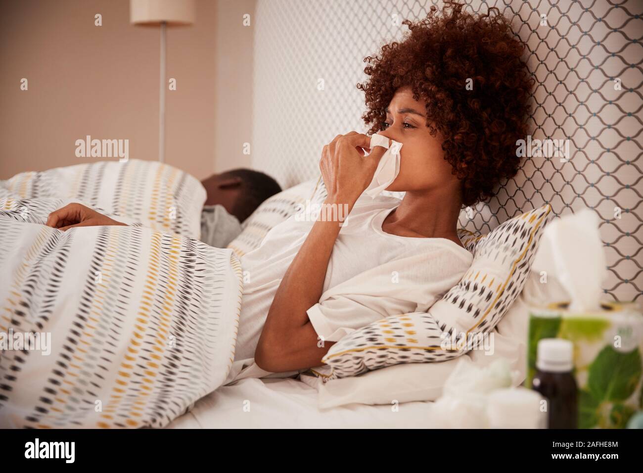 La millenaria americano africano donna seduta sul letto soffia il naso, partner addormentato in background, close up Foto Stock