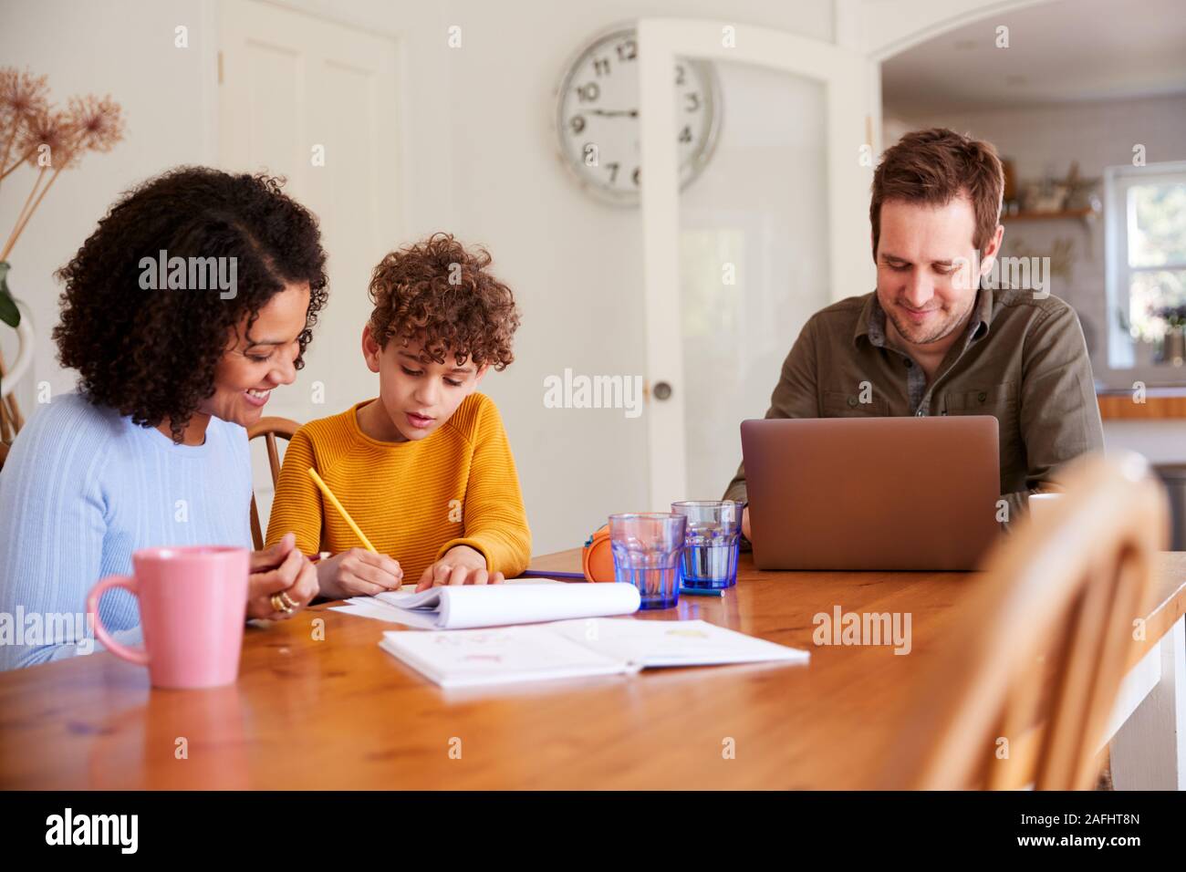Padre lavora sul laptop come madre figlio aiuta con i compiti sul tavolo da cucina Foto Stock