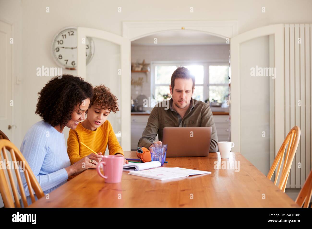 Padre lavora sul laptop come madre figlio aiuta con i compiti sul tavolo da cucina Foto Stock