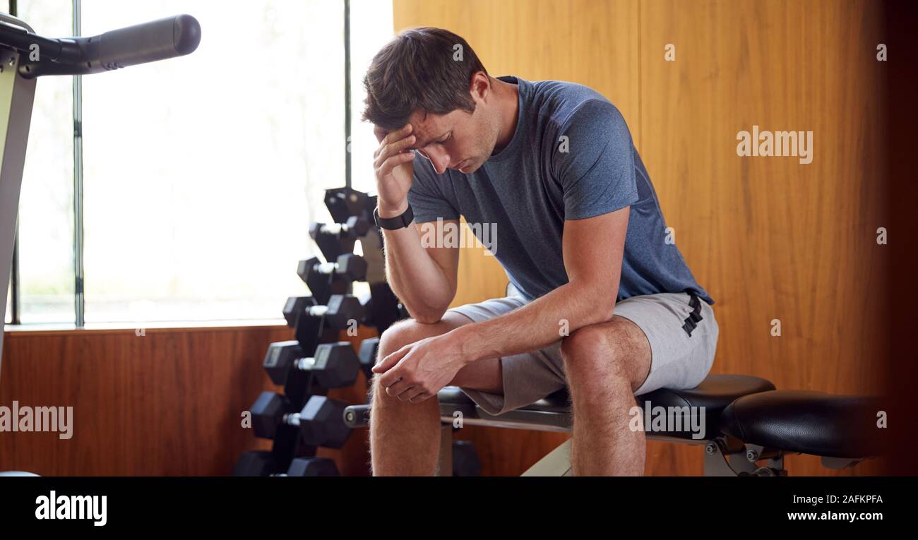 L'uomo preoccupato per l'immagine del corpo seduto sulla panca pesi nella palestra di casa Foto Stock