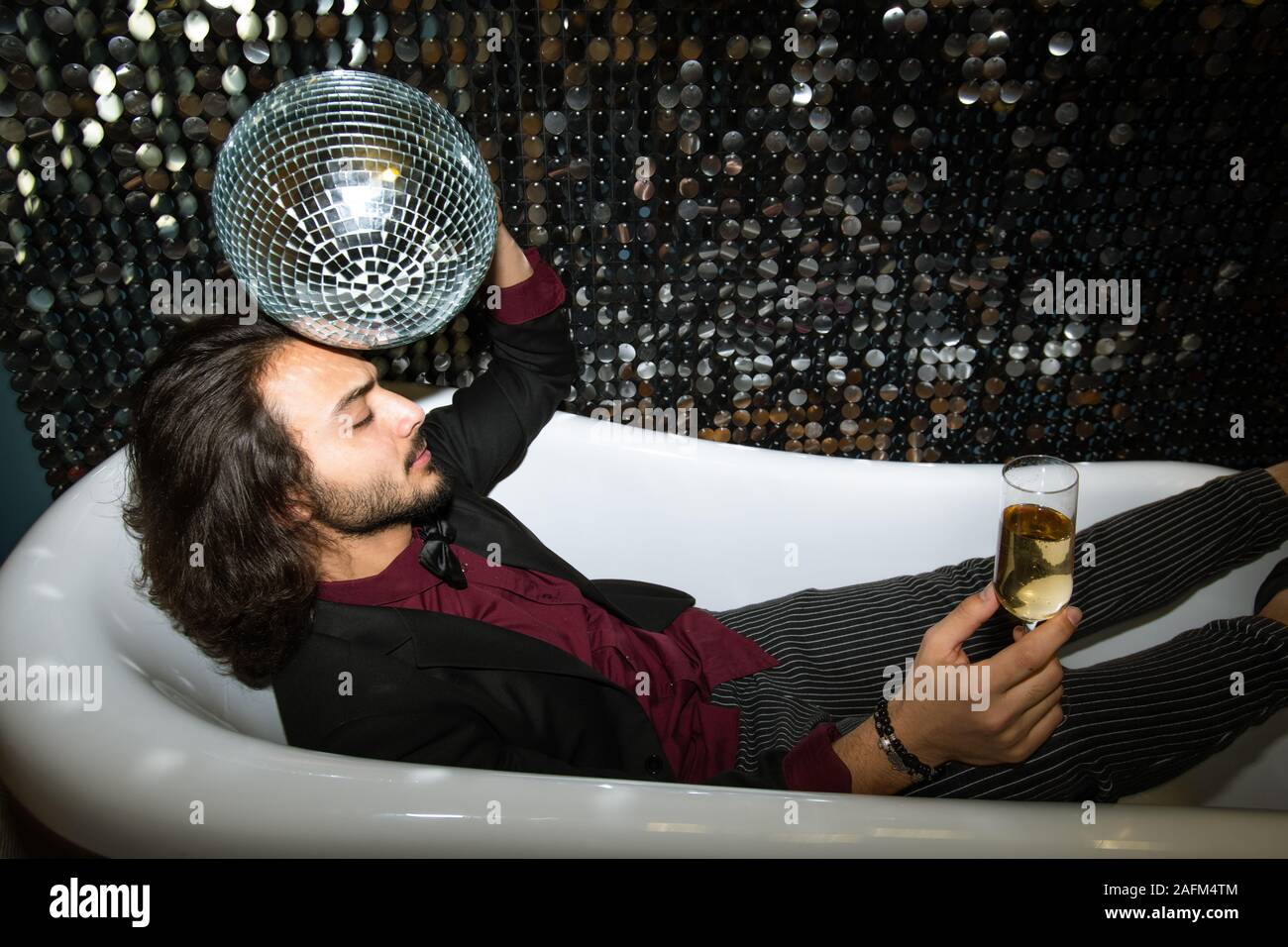 Stanco uomo con palla da discoteca dalla testa e flûte di champagne relax nella vasca da bagno Foto Stock