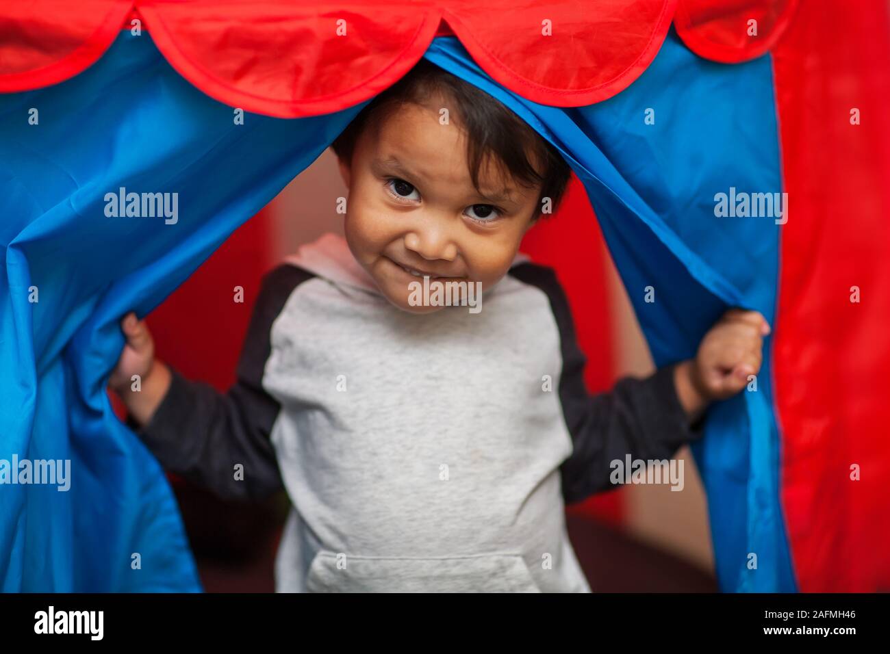Un giovane ragazzo di aprire le tende ad un rosso e blu tenda play e in piedi all'ingresso con un allegramente malizioso sorriso. Foto Stock