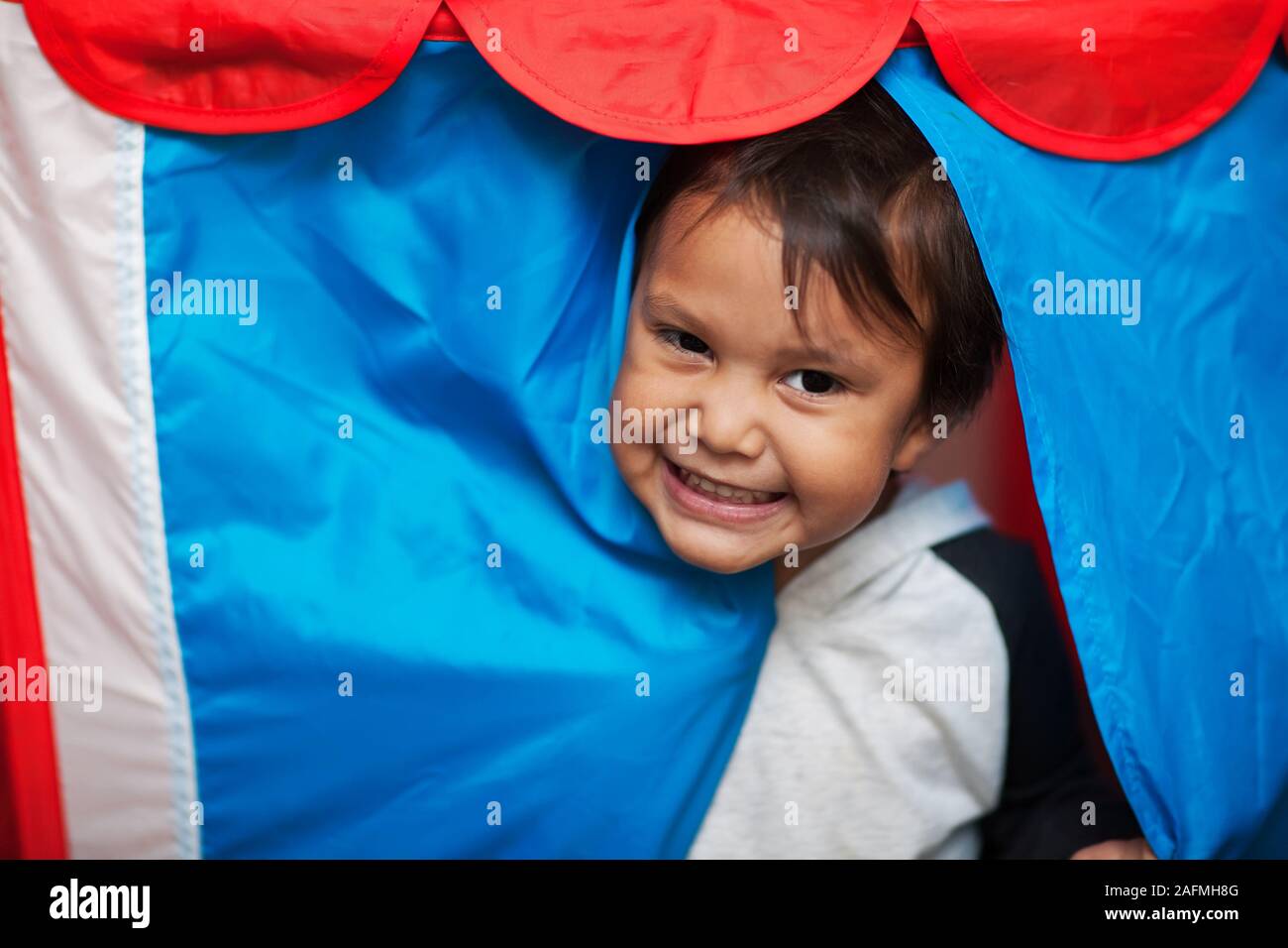 Carino piccolo ragazzo che è bloccata la sua testa fuori di un colorato gioco tenda e sorridente. Foto Stock