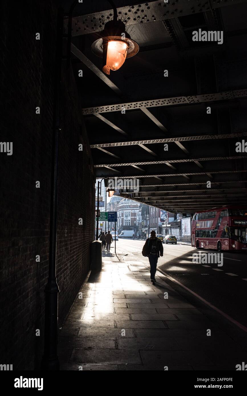 Scena di strada in Shoreditch, East London con ombra silhouette di una persona a piedi sotto un ponte stradale e red London bus Foto Stock