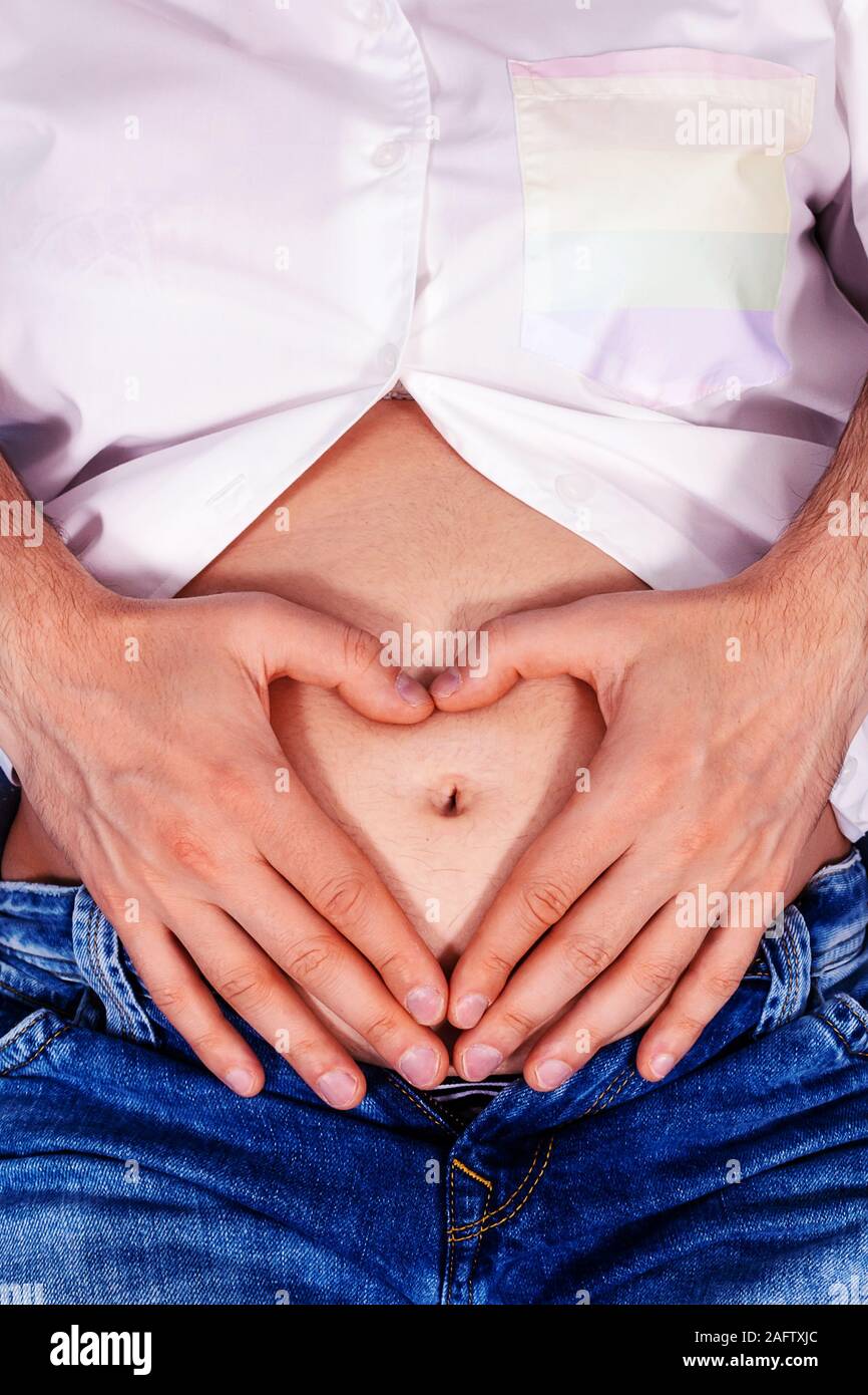 Transgender tiene per mano a forma di cuore sulla pancia in gravidanza. Baby concepimento in vitro. L'inseminazione artificiale. LGBTQ colori sulla tasca della camicia. Foto Stock
