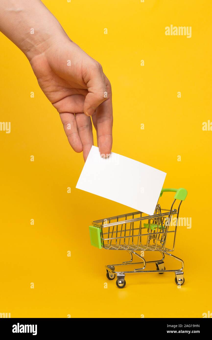 Mano maschio preso il banner bianco con spazio di copia da un piccolo carrello di metallo da un supermercato su sfondo giallo. Posto per il testo. Concetto di commercio. Foto Stock