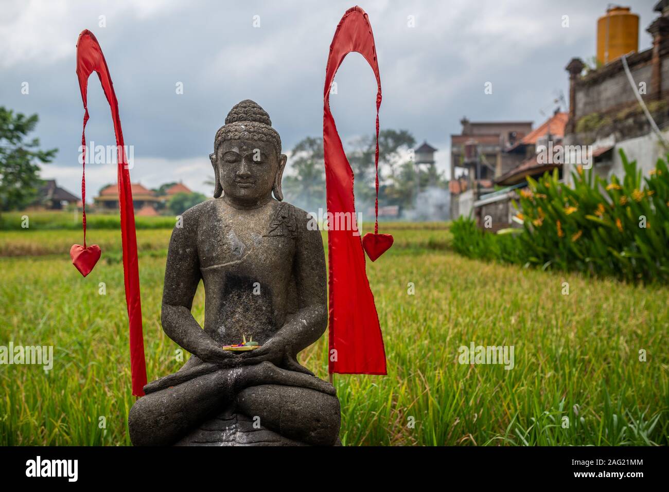 Statua di Buddha con bandiere rosse e sfondo sfocato di campi di riso, presa su un pomeriggio nuvoloso, Ubud, Bali, Indonesia Foto Stock