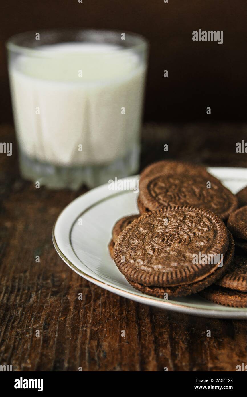 Colpo di primo piano verticale di un piatto di Oreos ripieni di cioccolato e un bicchiere di latte sullo sfondo Foto Stock