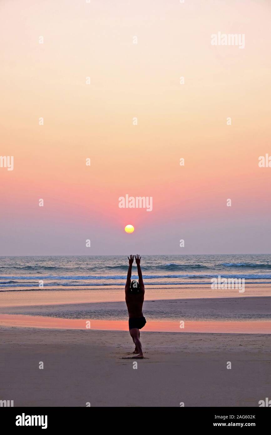 Un irriconoscibile uomo facendo un po' di yoga asana saluto al sole su di una vasta apertura vuota spiaggia sabbiosa, egli è la piegatura con braccia tese fino al sole, Foto Stock