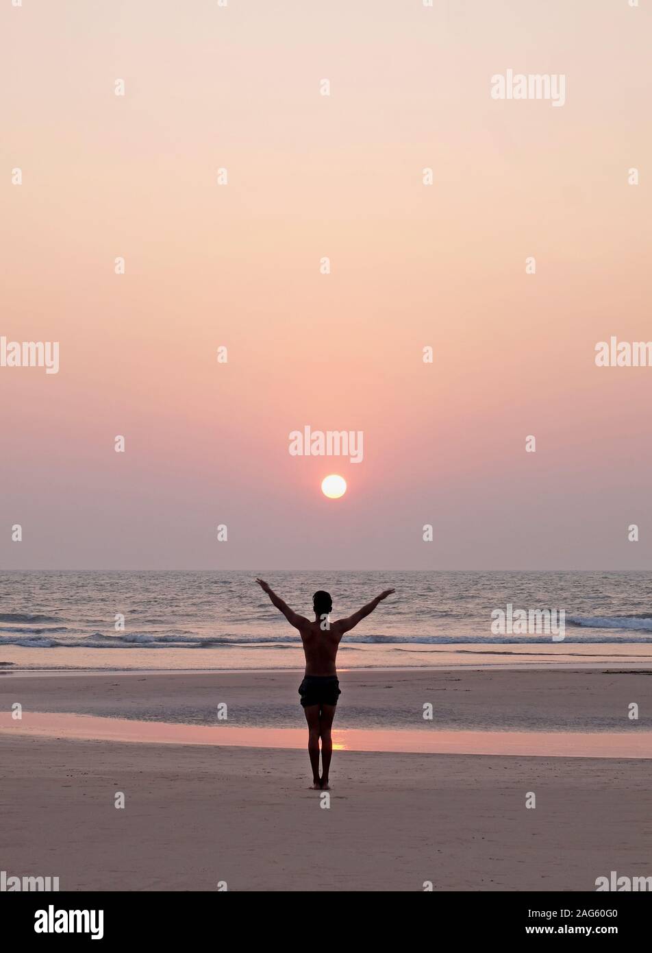 Un irriconoscibile uomo facendo un po' di yoga asana saluto al sole su di una vasta apertura vuota spiaggia sabbiosa, egli è in piedi con le braccia aperte, egli è rivestito u Foto Stock