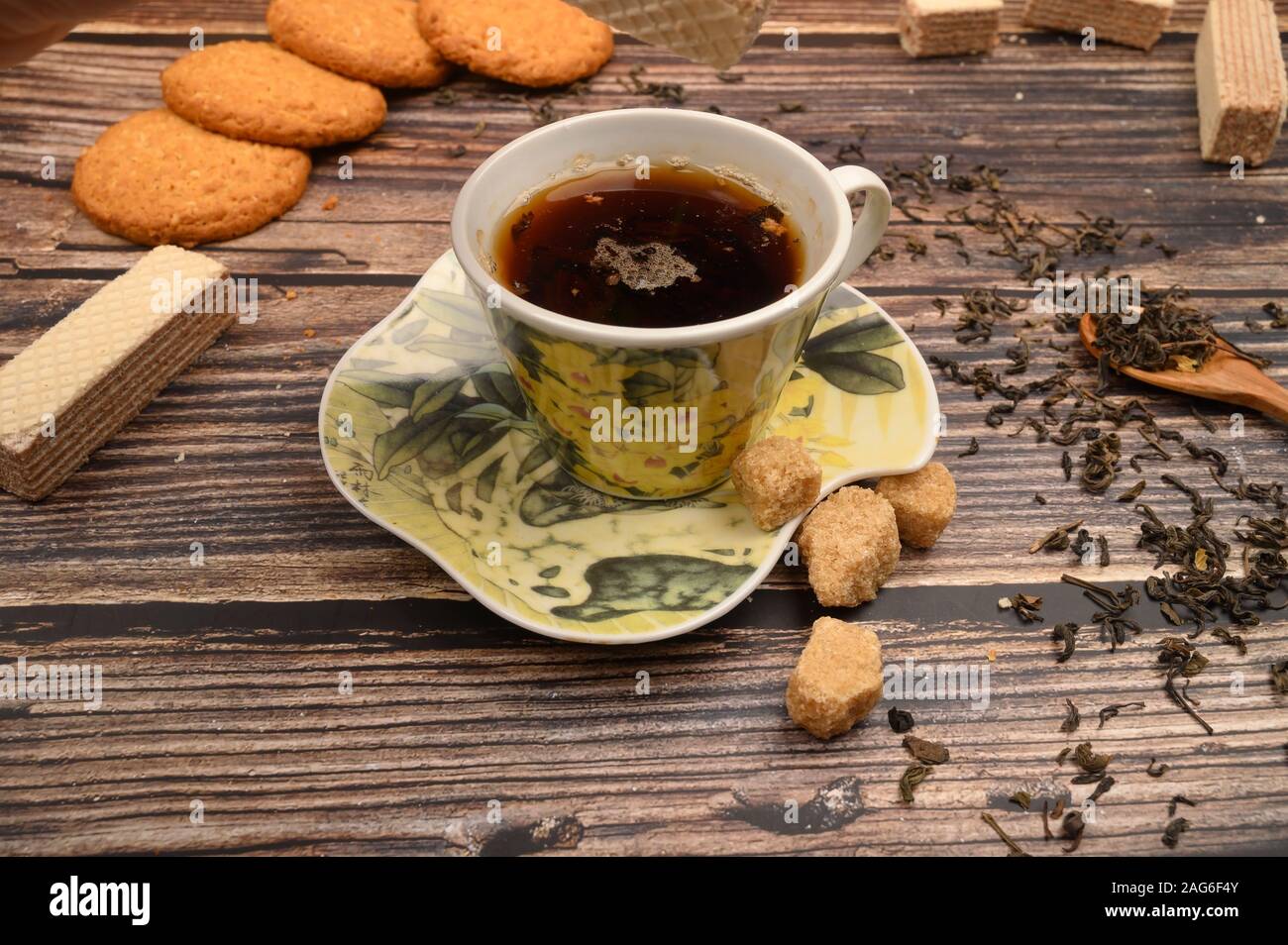 Una tazza di tè nero, foglie di tè, pezzi di zucchero di canna, fiocchi d'avena biscotti, cialde su uno sfondo di legno. Close up Foto Stock
