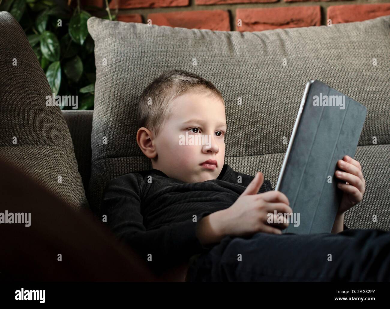 Ragazzo seduto sul divano nel soggiorno e guardare cartoni sul tablet. Ritratto di una smart pre-scuola per bambini utilizzando dispositivi a casa. Moderno kid. Foto Stock