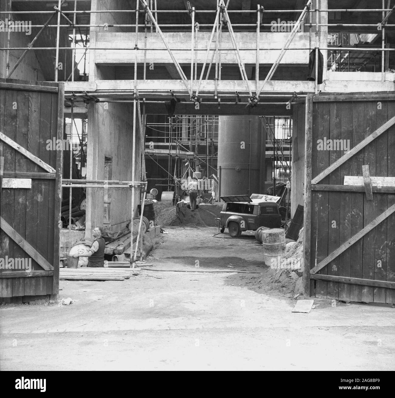 1960, storico, una vista attraverso le porte d'ingresso aperte in legno che mostrano l'interno di un cantiere, con impalcature e colonne di fondazione in cemento, Inghilterra, Regno Unito. Foto Stock