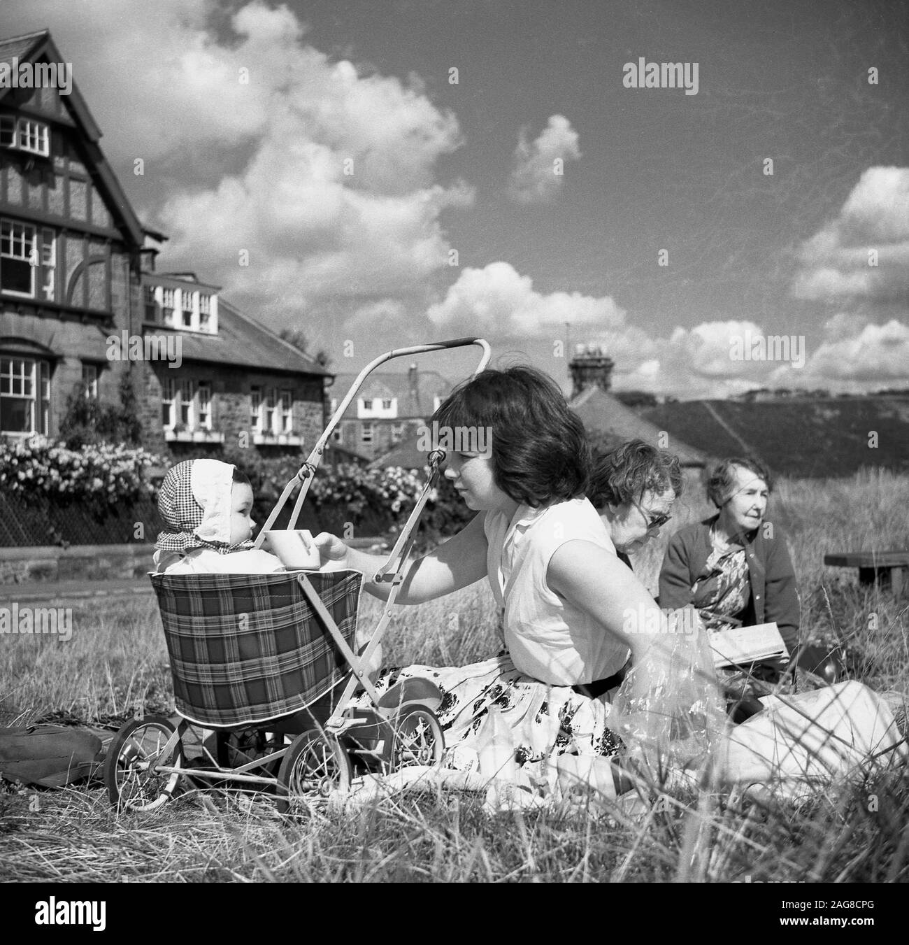 Anni '1960, storica, una madre e un bambino sedevano in un piccolo buggy o passeggino con un posto a sedere in tartan, seduti all'esterno su un'area erbosa alcune case, Inghilterra, Regno Unito. Foto Stock