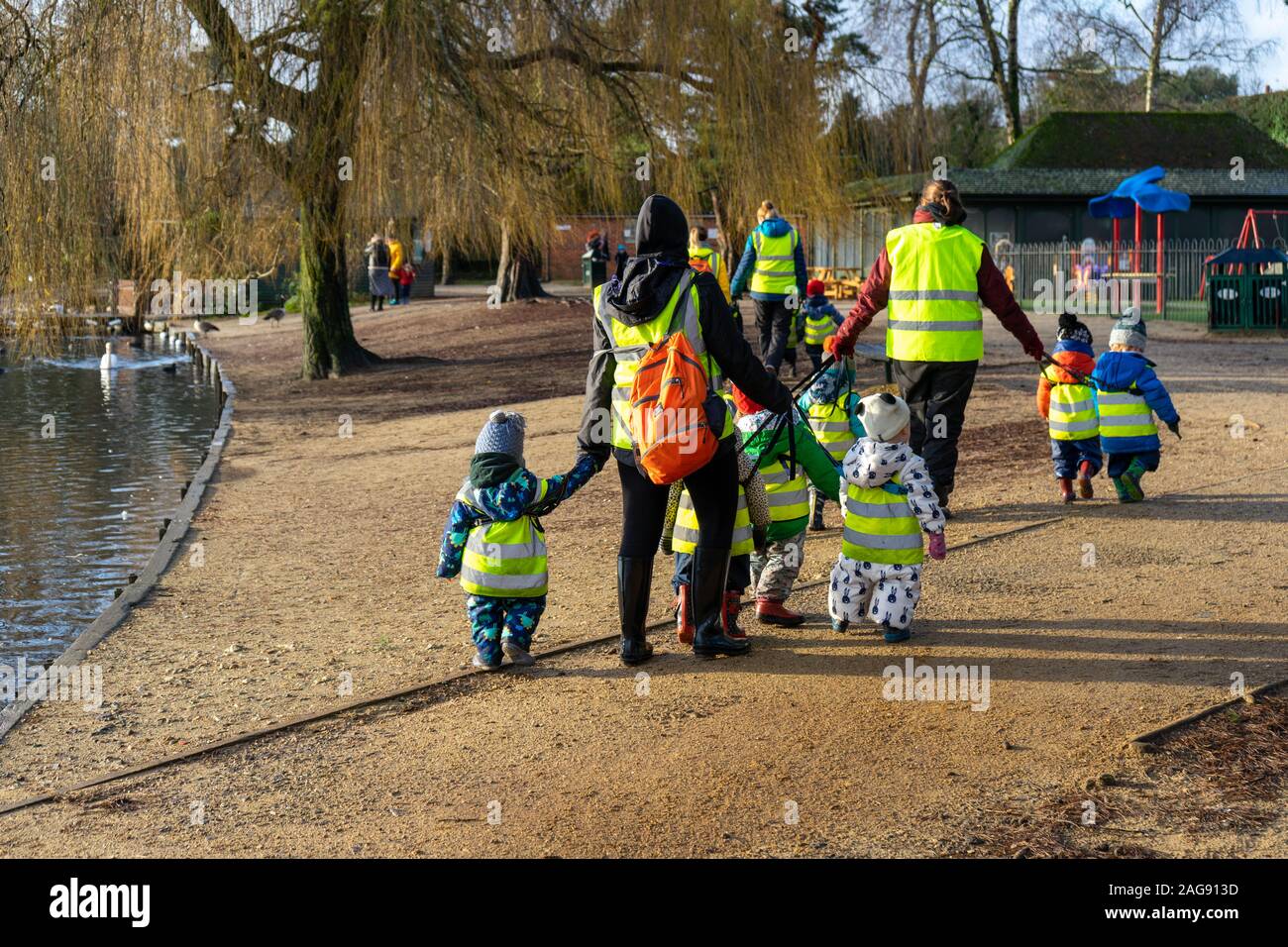 Un gruppo di bambini di una scuola materna in giacche ad alta visibilità a piedi con gli insegnanti o di servizi di custodia dei bambini lavoratori attraverso un parco in inverno Foto Stock