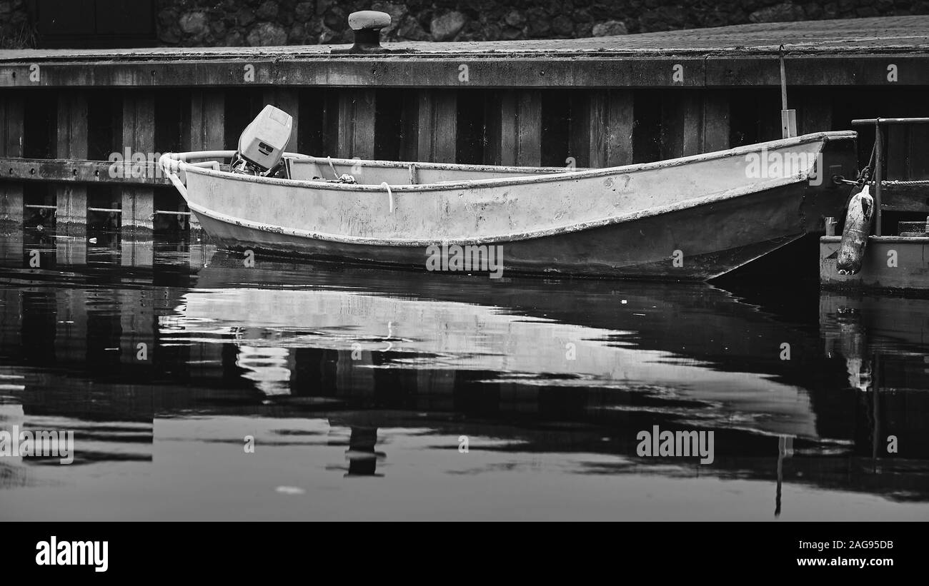 Immagine in scala di grigi di una barca arrugginita riflessa nel lago vicino a un molo Foto Stock