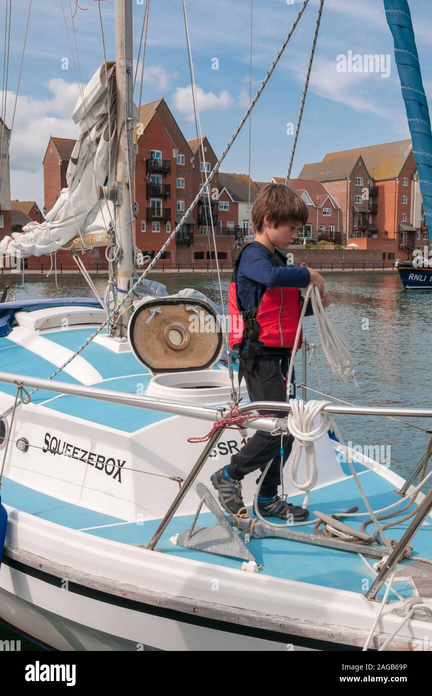 Ragazzo che indossa un giubbotto di salvataggio in piedi su un yacht, fune di bobinatura, Sovereign Harbour Marina, East Sussex, Regno Unito. Foto Stock