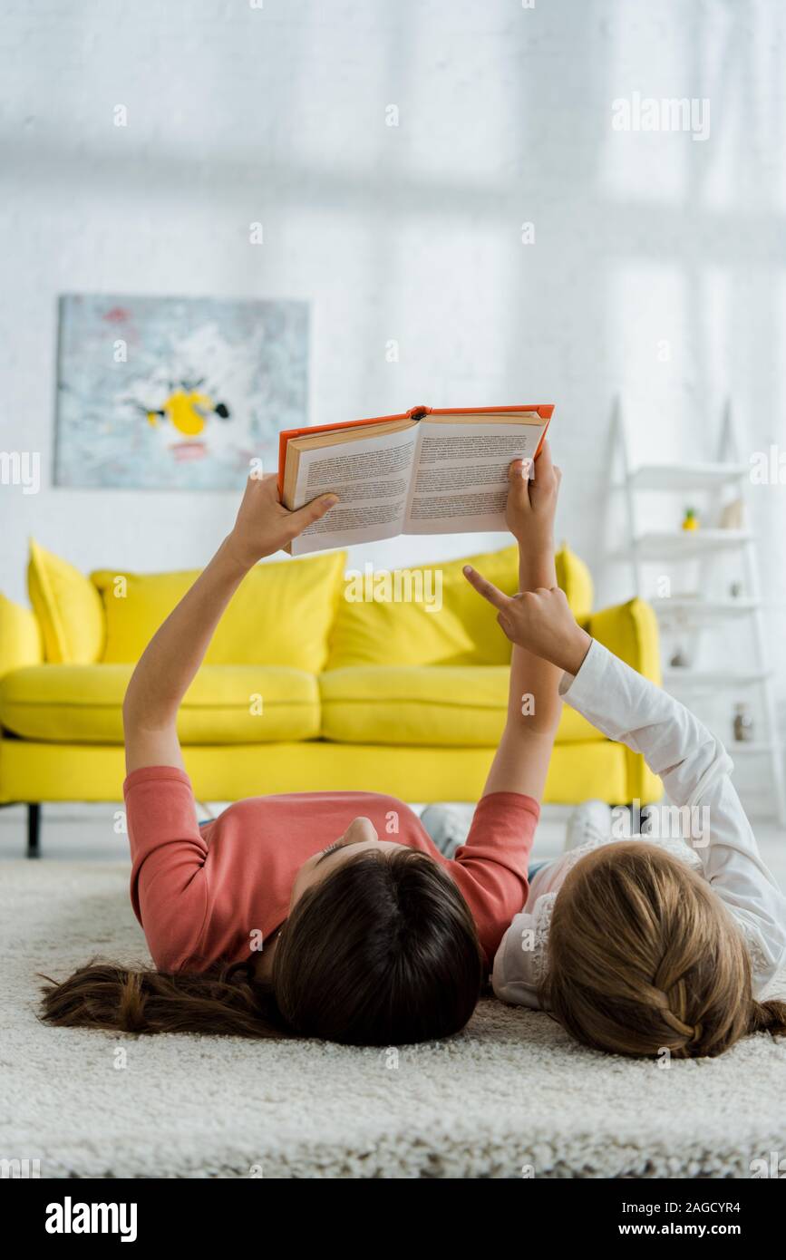 Bambino puntando con il dito sul libro mentre giaceva sul tappeto con servizio di babysitter Foto Stock