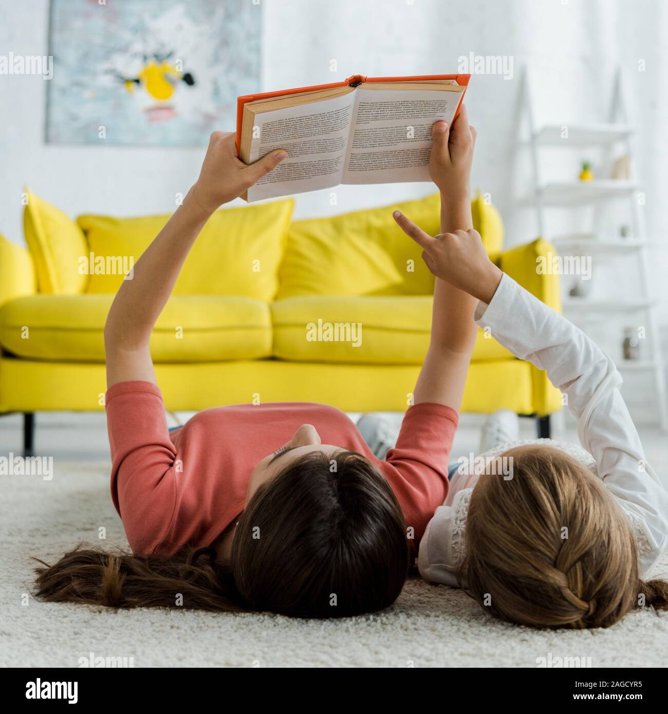 Kid puntando con il dito sul libro mentre giaceva sul tappeto con servizio di babysitter Foto Stock