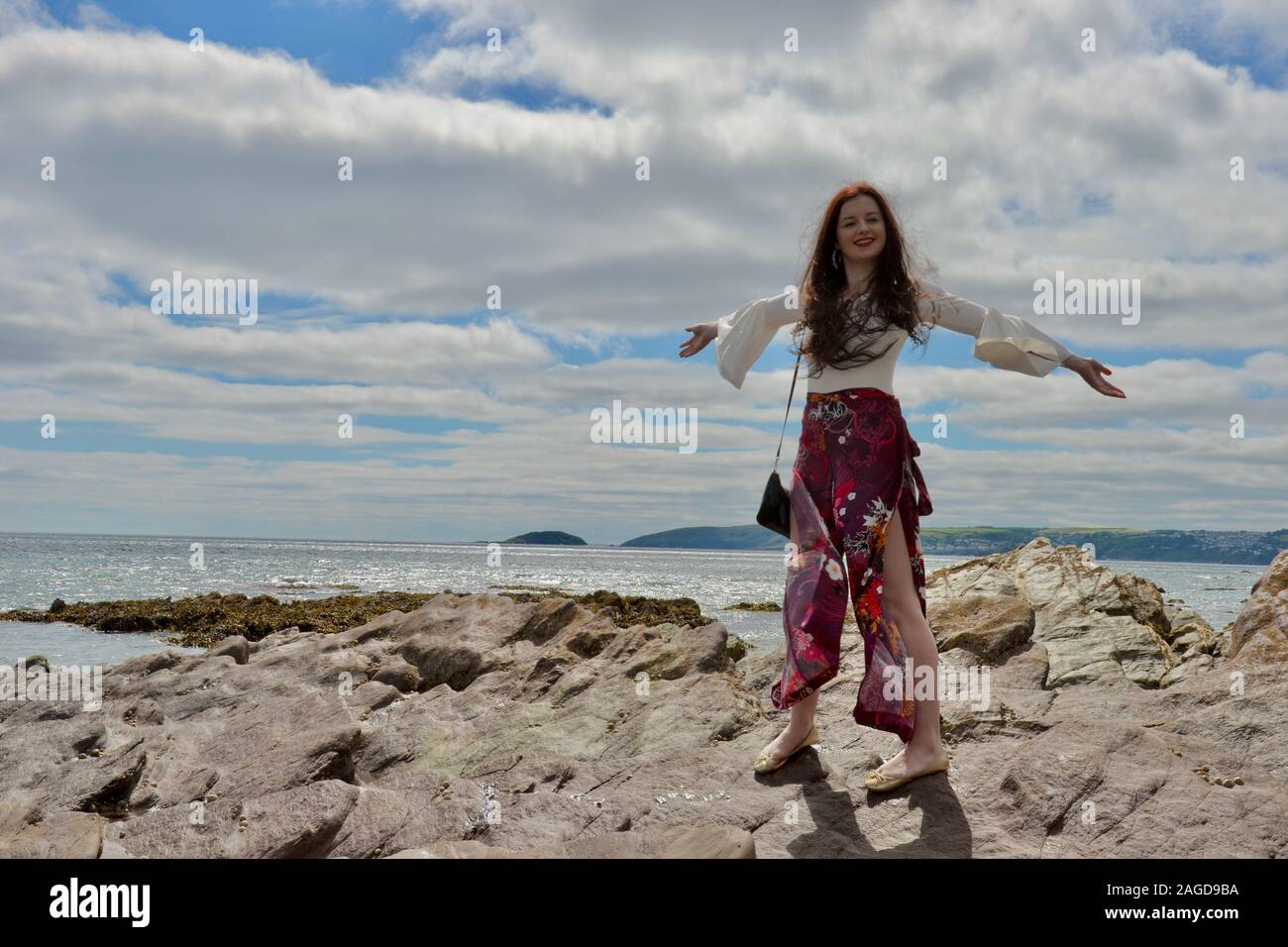 Hippie di moda giovane donna con capelli lunghi marrone e floreali pantaloni svasati in posa da mare sulle rocce sotto una cloudscape, braccia e sorridente Foto Stock