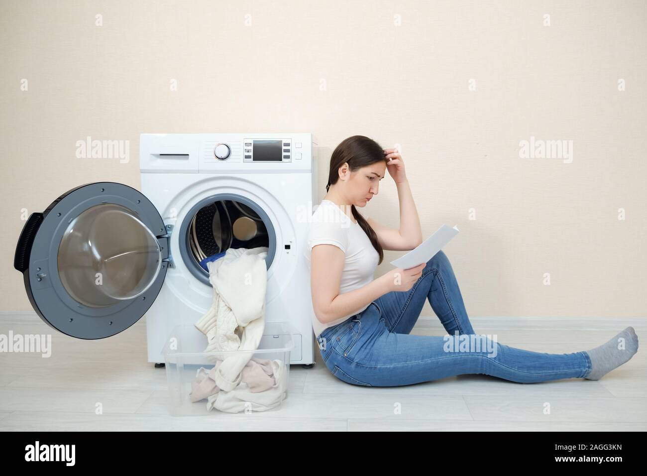 Sconvolto donna con biancheria sporca in scatola e manuale in mani si siede sul pavimento da una moderna macchina di lavaggio in camera Foto Stock