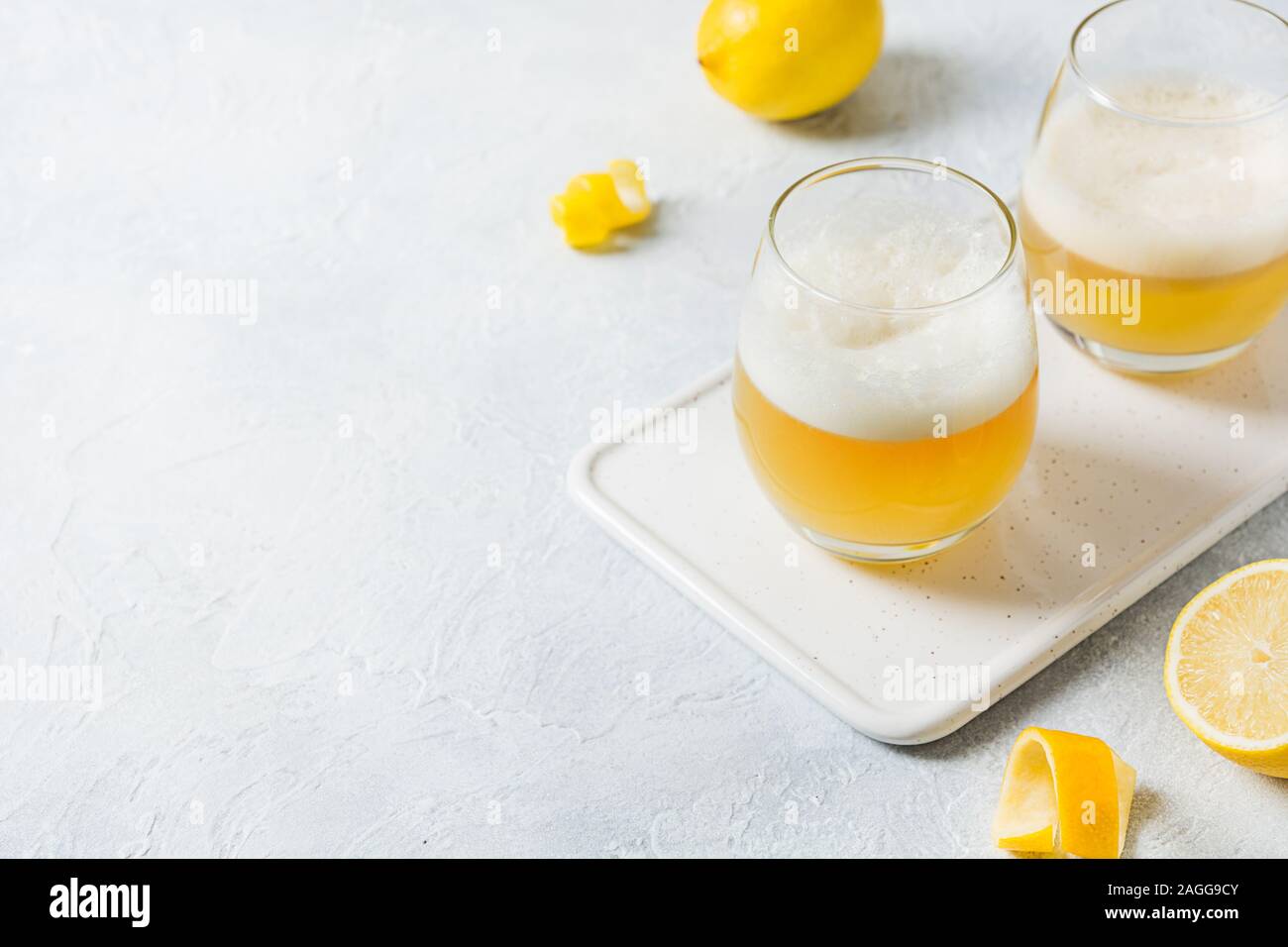 Due whiskey sour cocktail con gli ingredienti, il succo di limone e lo sciroppo di zucchero e albume in vetro sul tavolo bianco. Orientamento orizzontale. Foto Stock