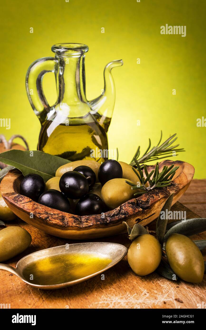 Su tavola in legno rustico, un intero cucchiaio di olio una ciotola di legno con olive e sullo sfondo di un ampolla Foto Stock