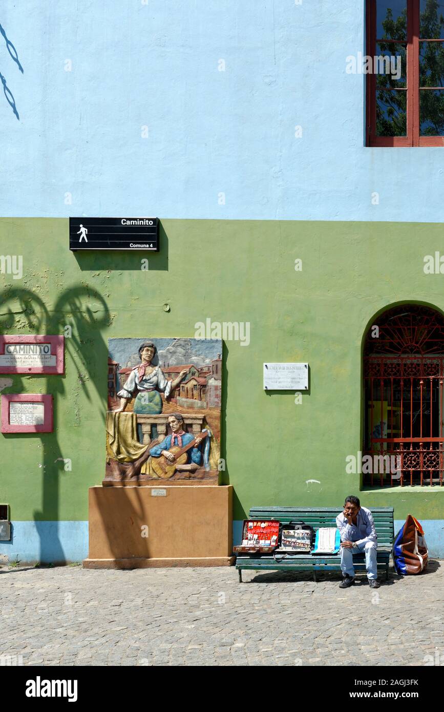 Street hawker con nessun cliente seduto su una panchina da un murale in colorate Caminito, Buenos Aries Foto Stock