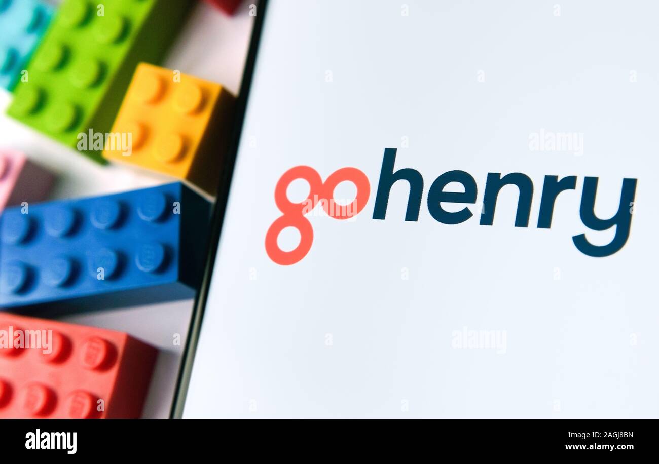 Logo GoHenry sullo schermo dello smartphone e alcune banconote e pezzi di lego su uno sfondo sfocato. Gohenry è una nuova carta prepagata per i bambini. Foto Stock