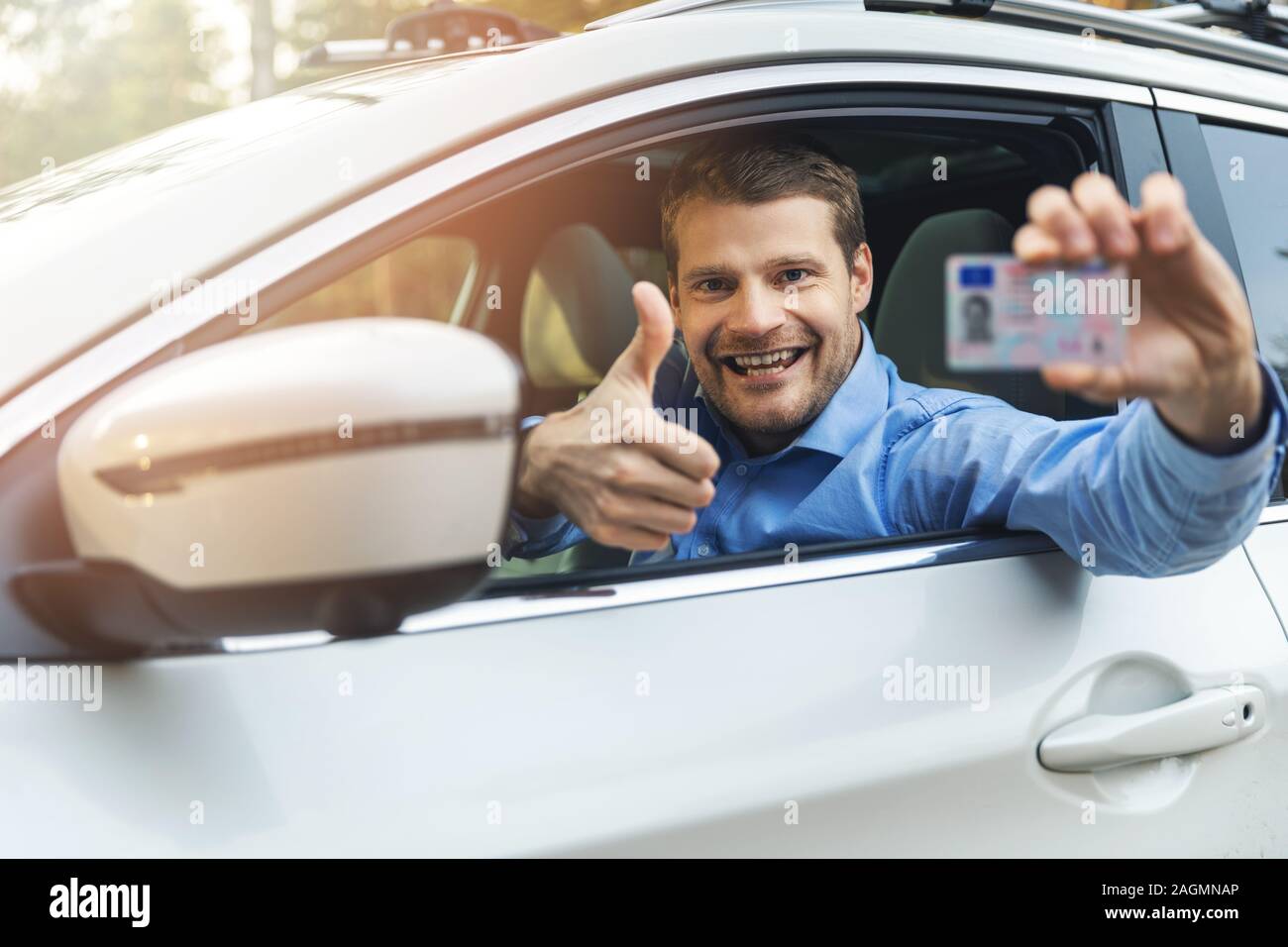 Giovane uomo sorridente seduto in macchina e che mostra la sua nuova licenza di pilota con il pollice in alto segno al di fuori della finestra di auto Foto Stock