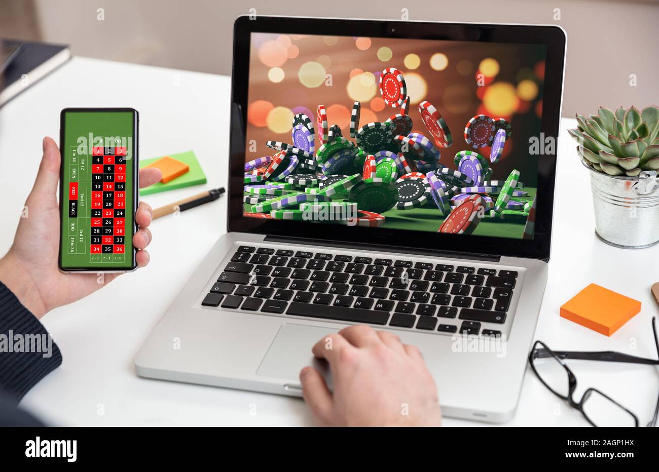 Online gioco d'azzardo, scommesse concetto. Poker chips, casinò roulette su laptop e schermate Telefono. Uomo che lavora con un computer e tenendo uno smartphone, offi Foto Stock