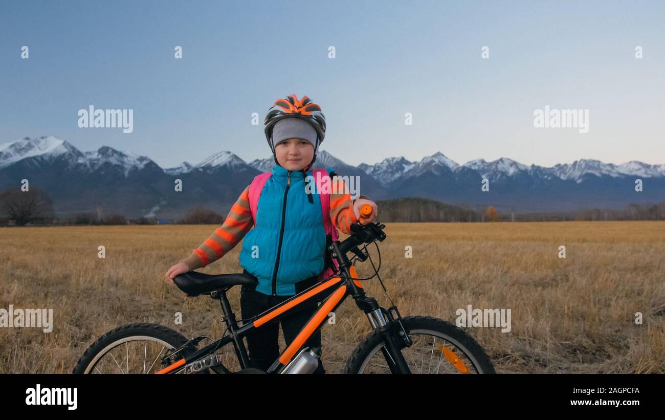 Uno dei bambini caucasici a piedi con bici nel campo di grano. Poco ragazza camminare nero ciclo arancione su sfondo di belle montagne innevate. Foto Stock
