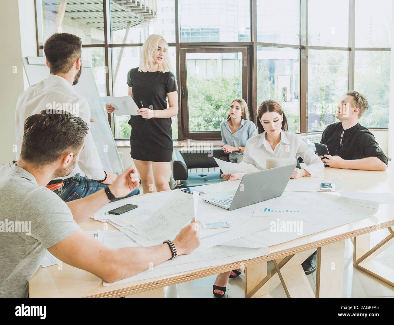 Una giovane donna in un abito nero parla di un progetto mentre in piedi vicino a una scheda dimostrativa. Il resto del personale stanno ascoltando con attenzione al suo. Foto Stock