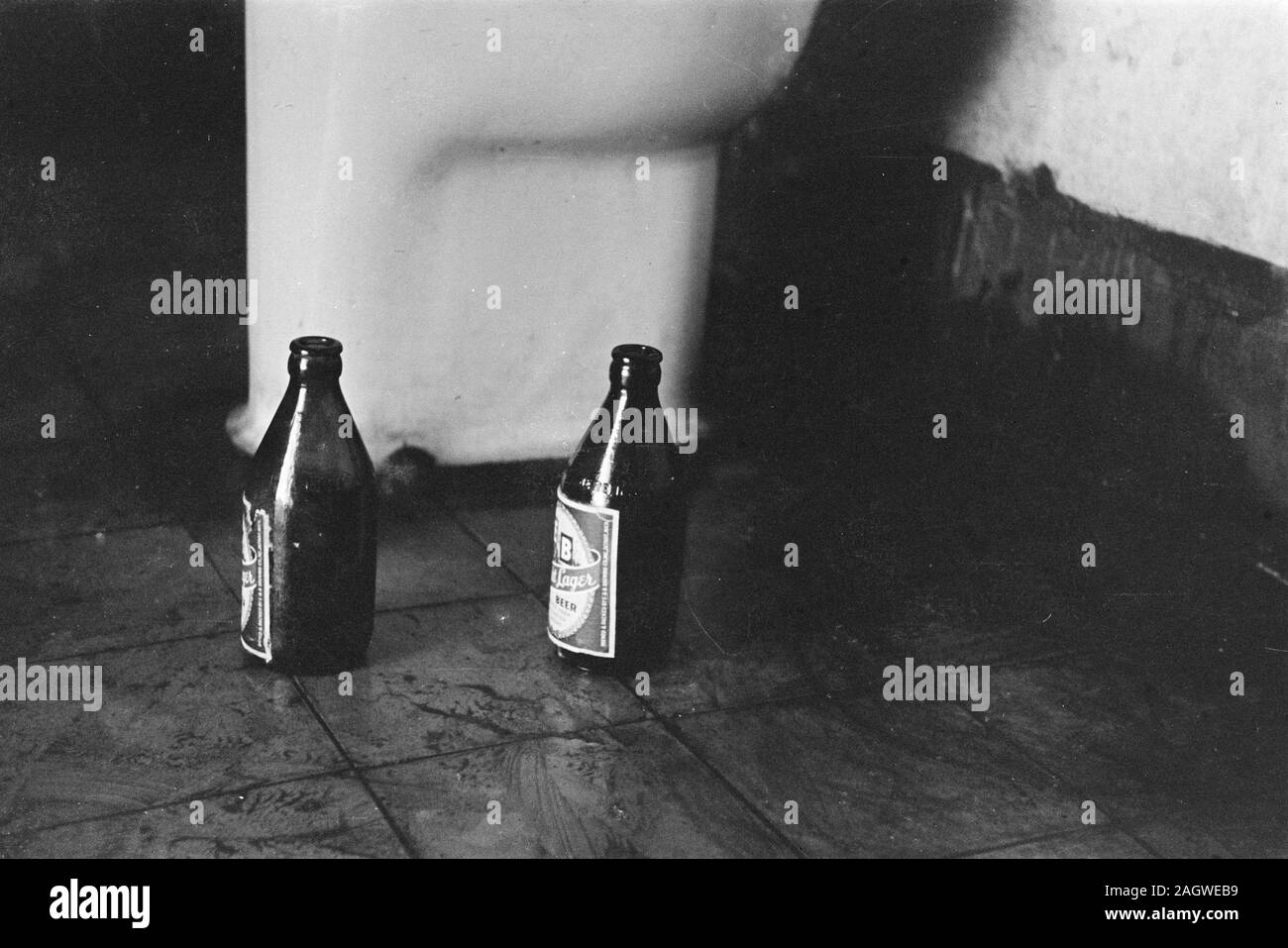 Due bottiglie di birra su di un pavimento di piastrelle in prossimità di una tazza di gabinetto - Marca: E&B luce birra lager; data 1947; posizione Indonesia, Indie orientali olandesi Foto Stock