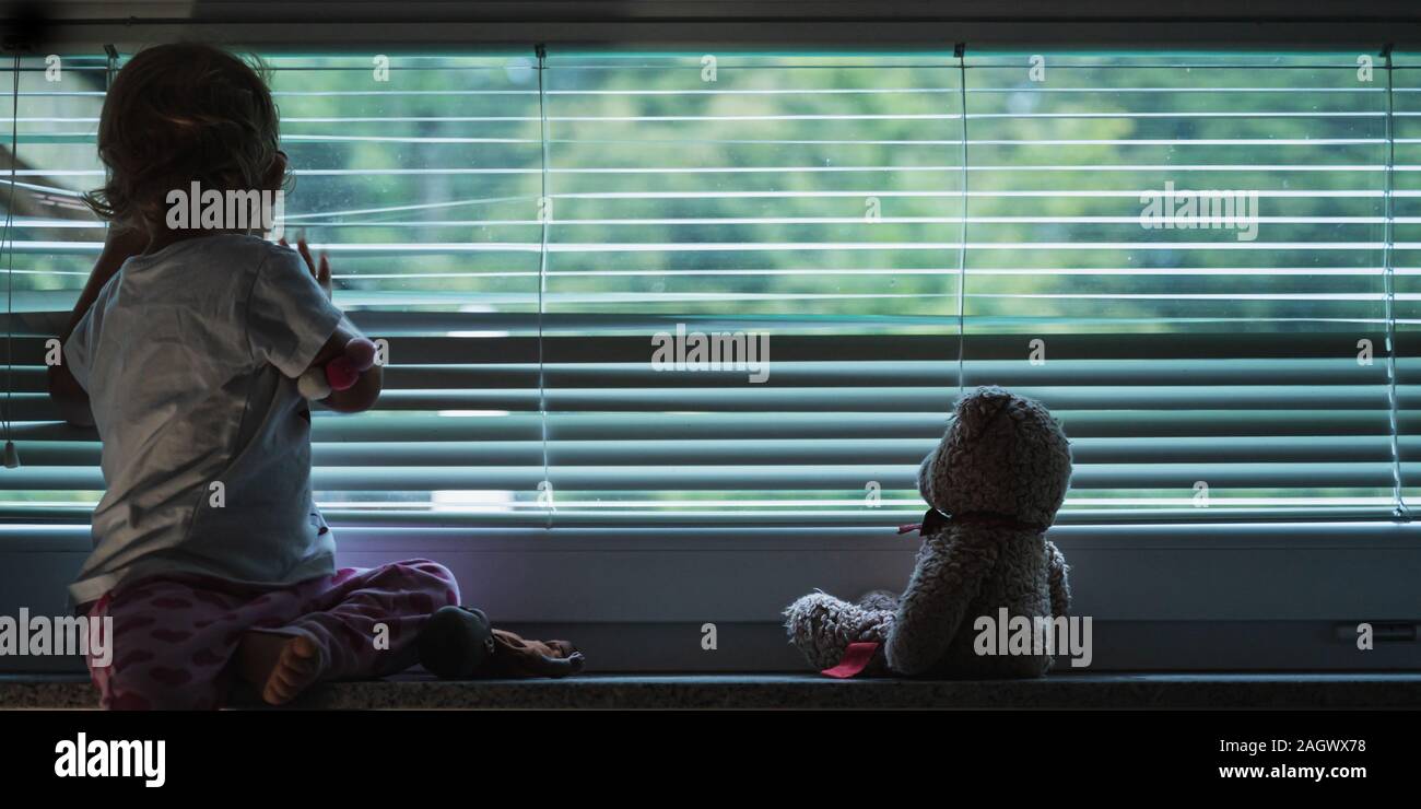 Il Toddler girl guardando fuori dalla finestra attraverso abbassato le tende alla veneziana con il suo orsacchiotto seduto accanto a lei in un'immagine concettuale. Foto Stock