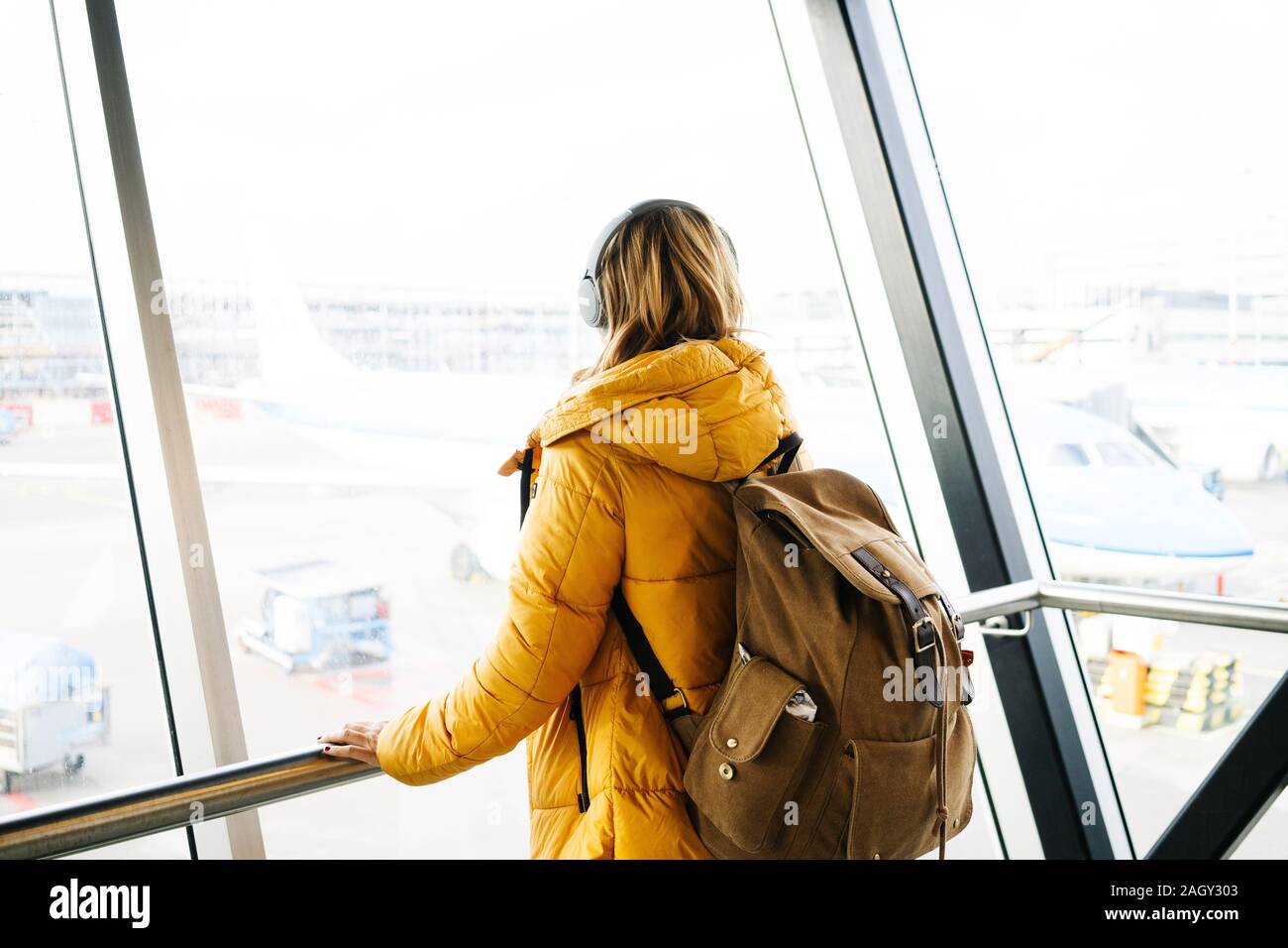 Una bionda caucasica donna cammina attraverso l'aeroporto con la giacca gialla e zaino Foto Stock