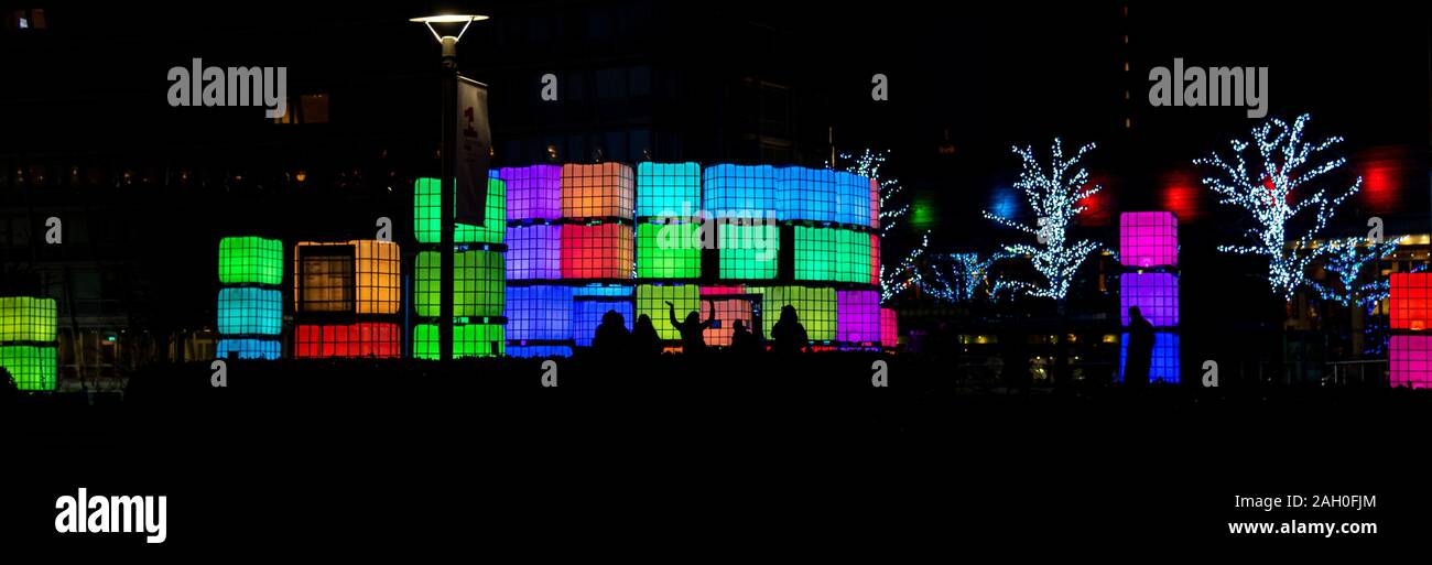 Vista panoramica di sagome di persone aventi il divertimento di notte mentre in posa di fronte dei cubetti illuminato con luce di diversi colori in Chavasse Park Foto Stock