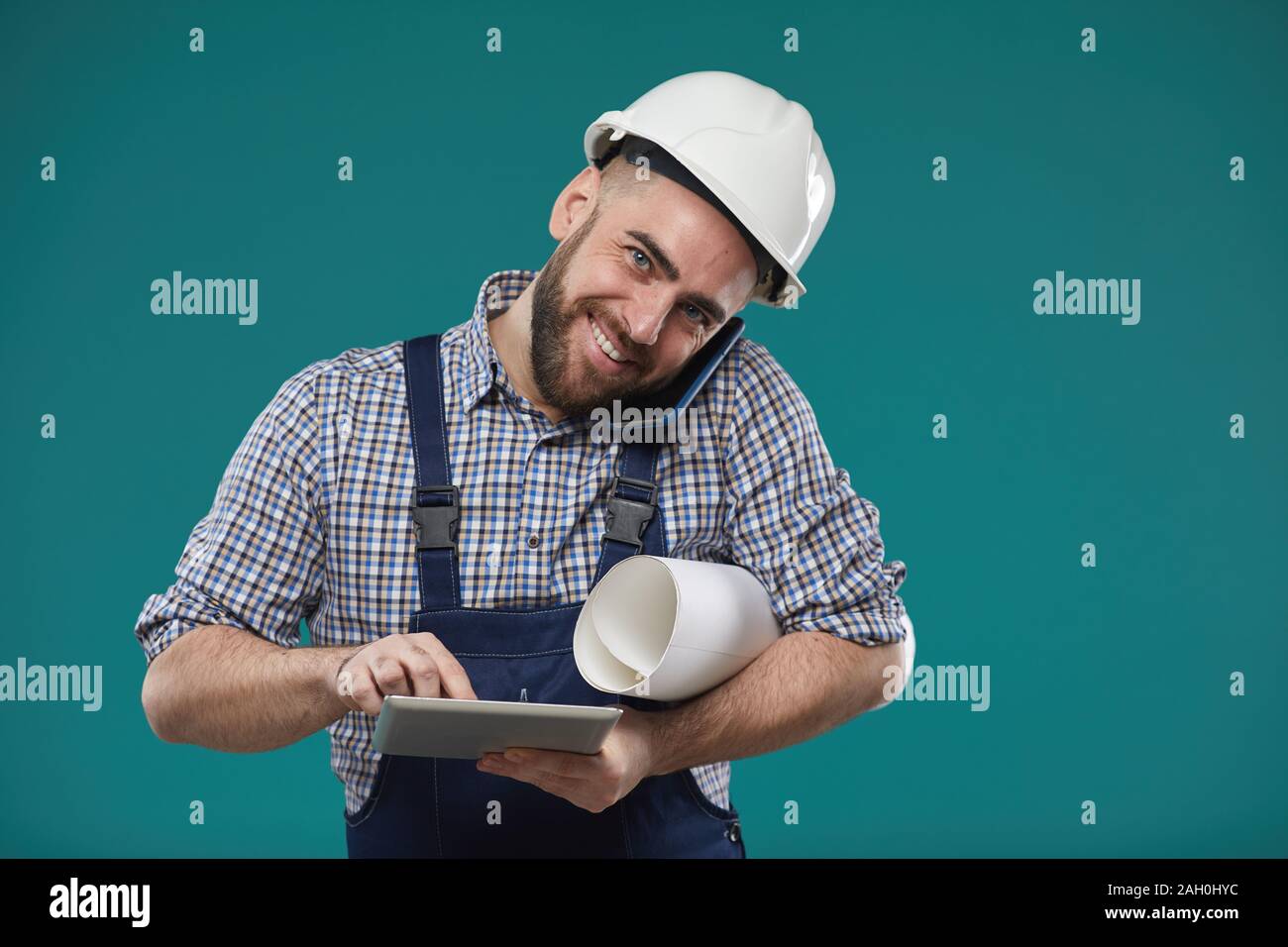 Orizzontale di studio shot della trafficata foreman azienda piano di costruzione utilizzando computer tablet e parlare al telefono allo stesso tempo Foto Stock