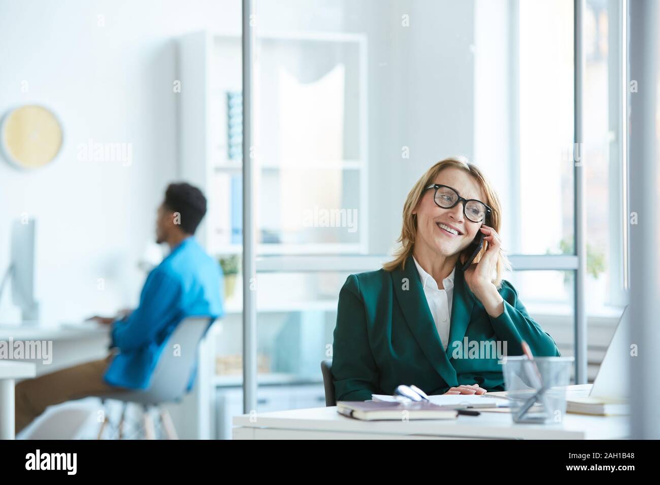 Coppia manager in occhiali seduta al tavolo parlando al telefono cellulare e sorridente in ufficio Foto Stock