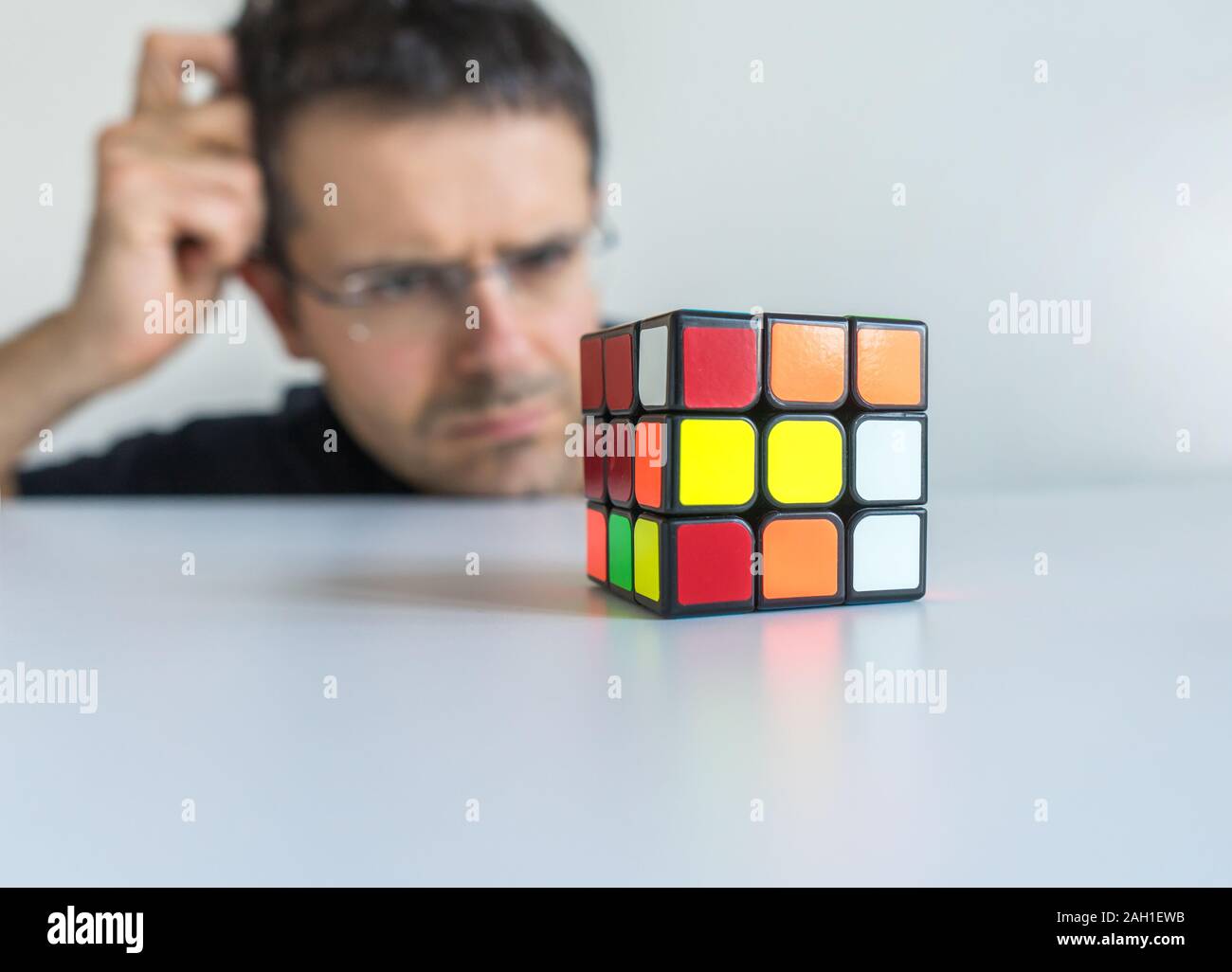 Uomo con occhiali guardando perplesso di fronte al cubo di Rubik graffiare la sua testa senza sapere come risolvere il problema Foto Stock