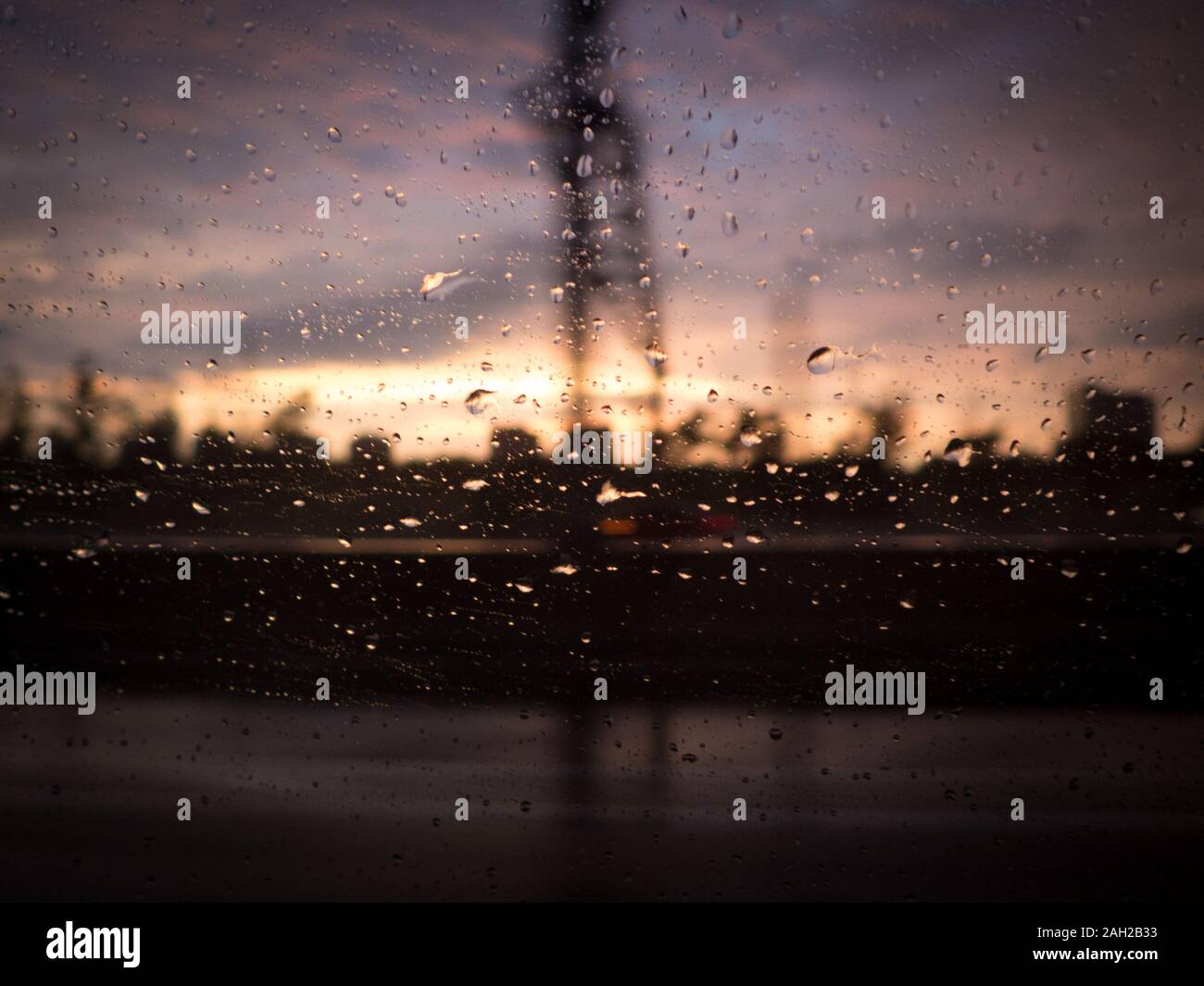 Silhouette sfocata di impostazione industriale attraverso il vetro per auto con gocce di pioggia contro la finestra. Tramonto e nuvole in background Foto Stock