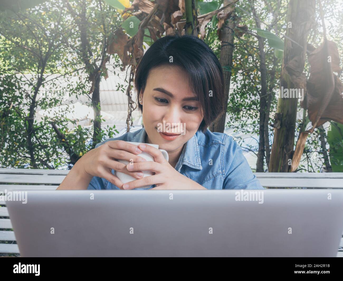 Felice bella donna asiatica di capelli corti jeans blu shirt bianco potabile tazza di caffè caldo e guardando sul computer portatile in cafe con verde bac Foto Stock