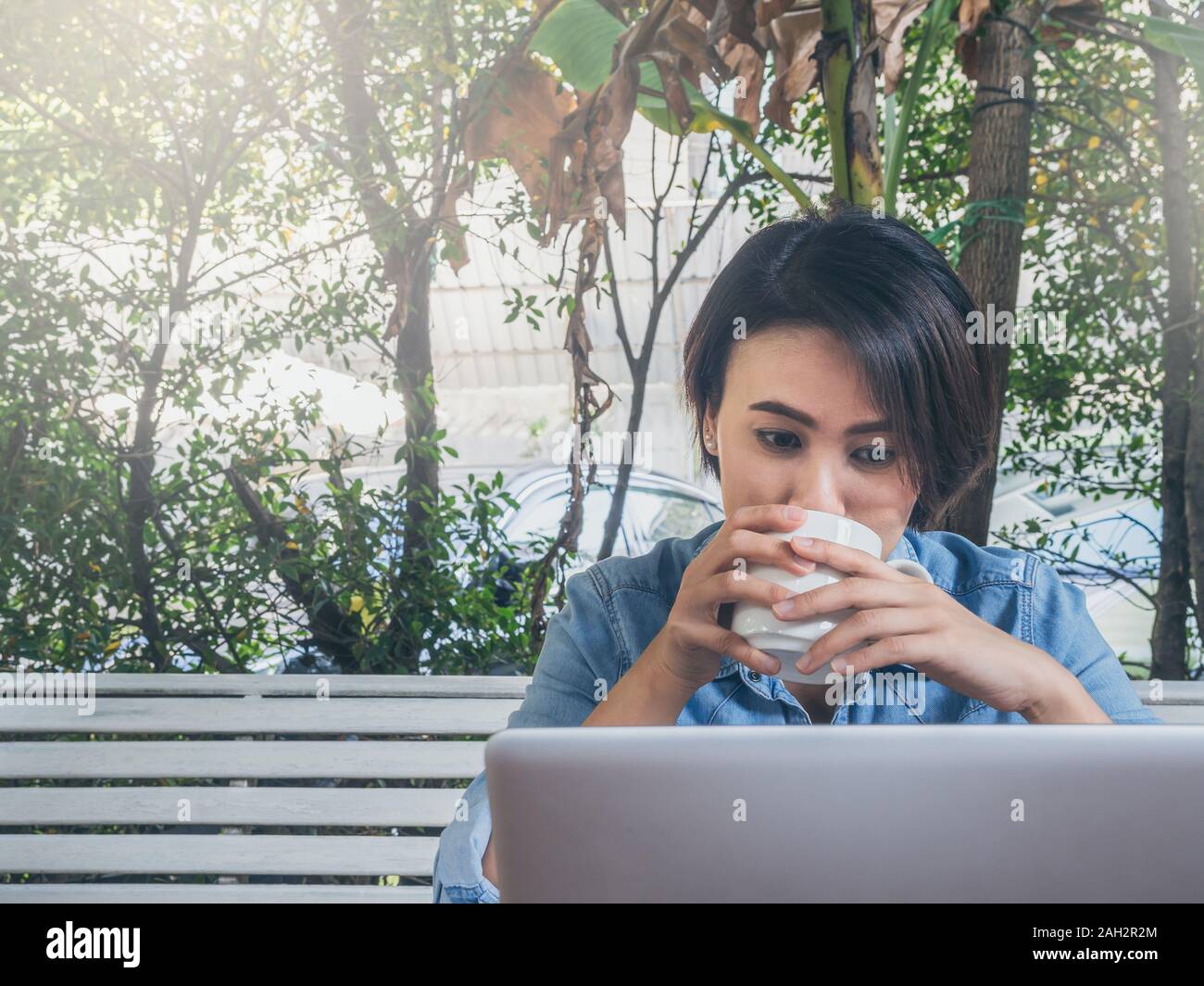 Felice bella donna asiatica di capelli corti jeans blu shirt bianco potabile tazza di caffè caldo e guardando sul computer portatile in cafe con verde bac Foto Stock