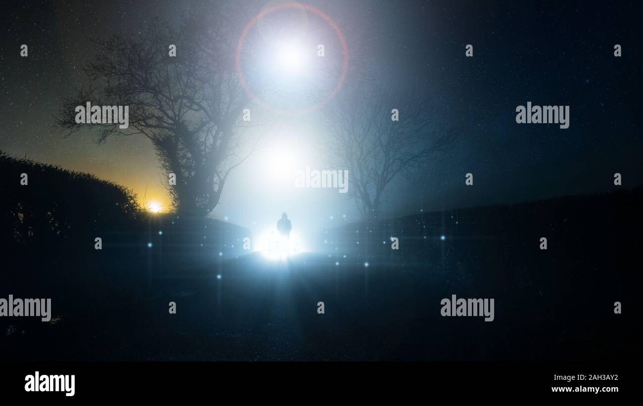Una soprannaturale, UFO concetto di una paurosa e misteriosa figura con cappuccio, in piedi di fronte ad un luminoso luci nel cielo, su un vicolo del paese, su un spooky fogg Foto Stock