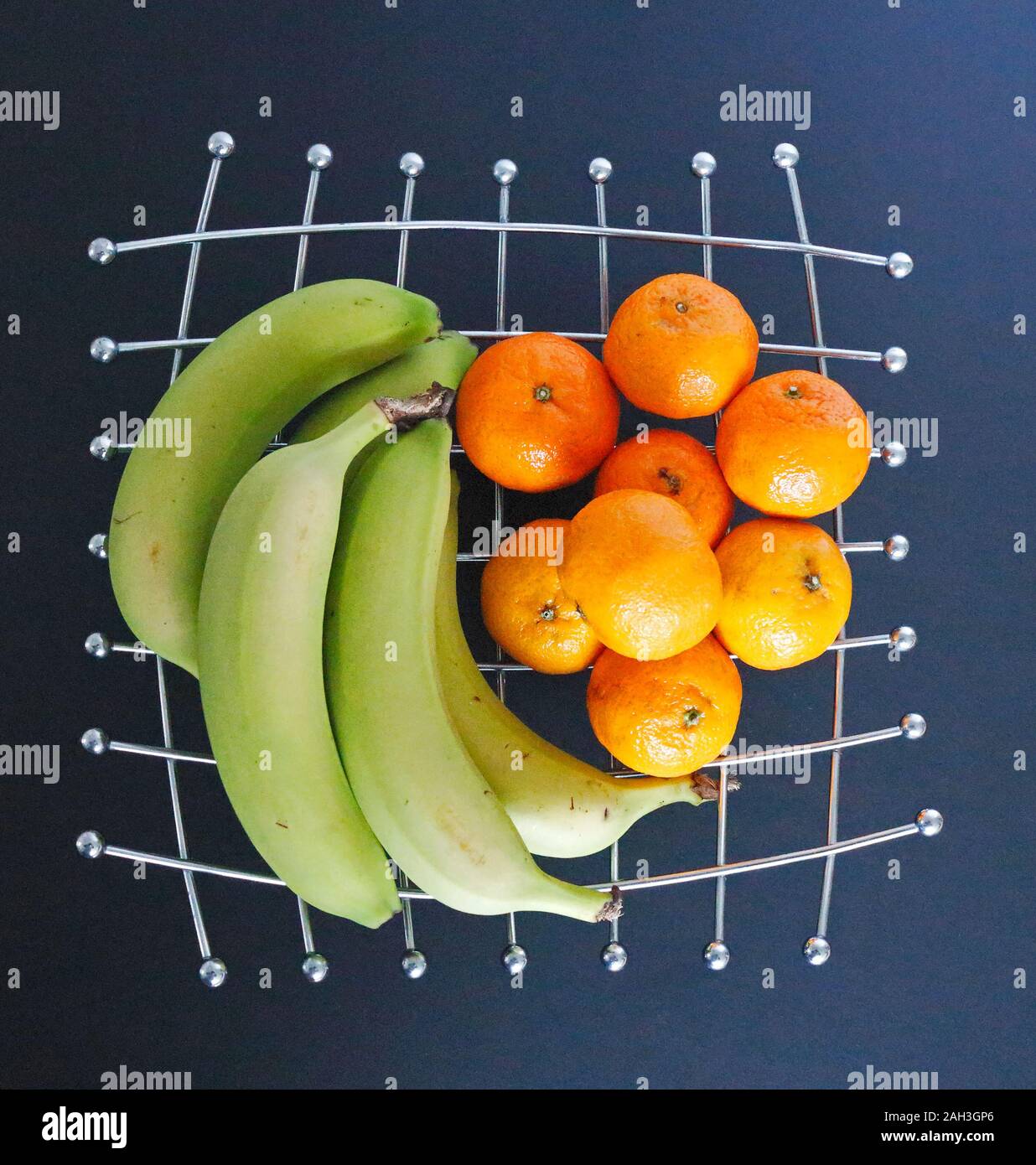 Un mazzetto di verde la maturazione delle banane e mandarini su un acciaio inossidabile rack di frutta con uno sfondo nero. Foto Stock