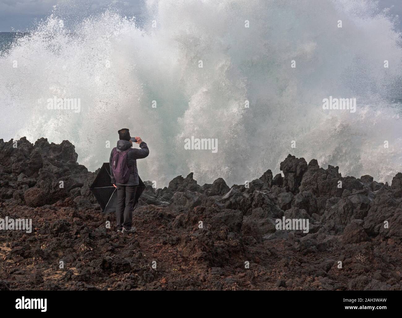 Una femmina di lone scatta una fotografia con un telefono cellulare di una grande onda che si schiantano contro le rocce. Lei sembra ignorare la posizione pericolosa ella è a. Foto Stock