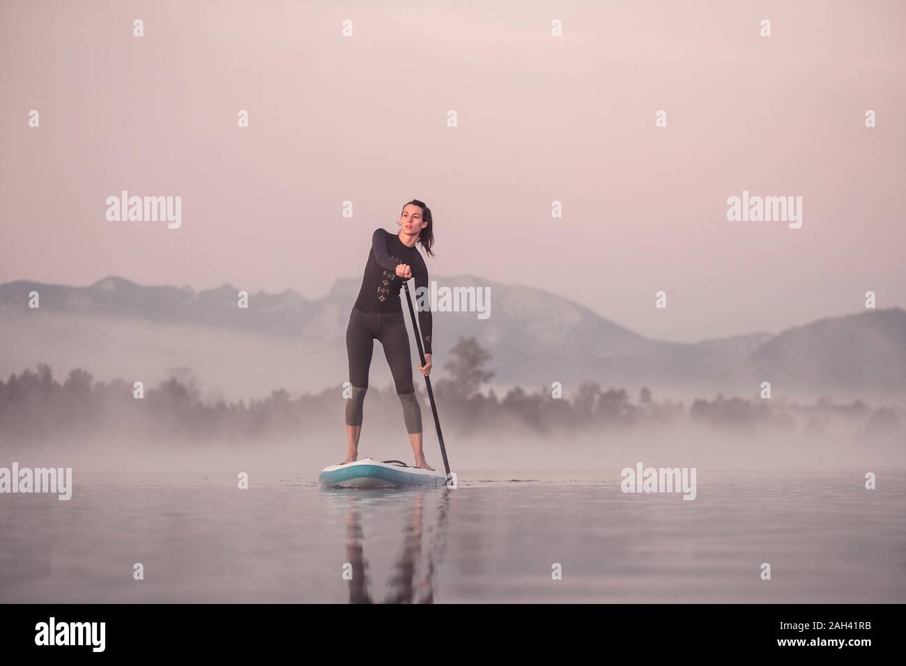 Donna stand up paddling sul lago Kirchsee presso il Morning mist, Bad Toelz, Baviera, Germania Foto Stock